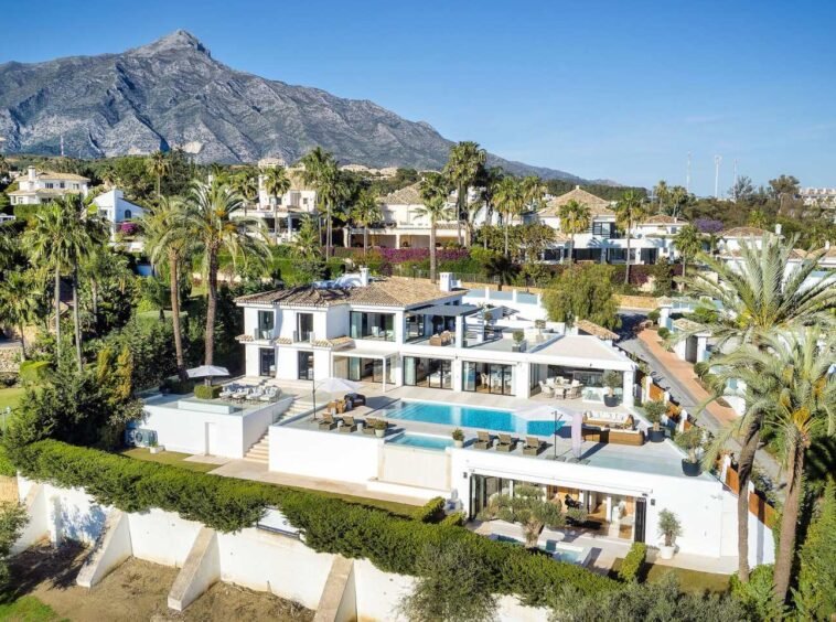 Stunning facade of a frontline golf villa in Nueva Andalucia, showcasing contemporary architecture with clean lines, large windows, and a spacious outdoor area. The villa is beautifully integrated into its lush green surroundings, offering a serene and luxurious atmosphere with views of the pristine golf course.