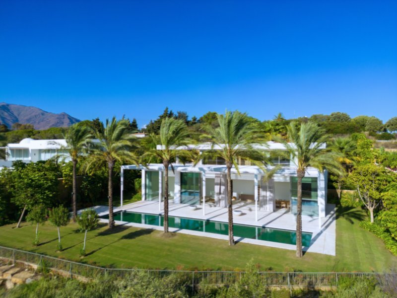 A striking modern villa with a sleek, contemporary facade, featuring clean lines, expansive glass windows, and a sophisticated design. The property is framed by lush landscaping, creating a perfect blend of luxury and nature. The villa's minimalist style is complemented by soft exterior lighting.