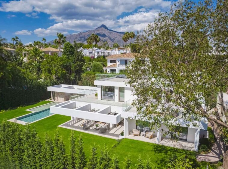 Stunning facade of a modern villa in Nueva Andalucía, featuring clean architectural lines, large windows, and a harmonious blend of sleek, contemporary design elements. The villa’s exterior is complemented by lush landscaping, creating an elegant and inviting first impression of luxurious living in a tranquil setting.