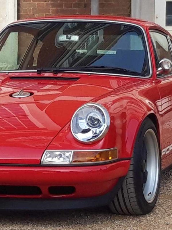 A custom Singer Porsche 911 parked on a scenic road, showcasing its classic design with modern refinements. The car features a sleek silver body, flared wheel arches, and distinctive retro-styled headlights, embodying the perfect blend of vintage elegance and high-performance engineering.